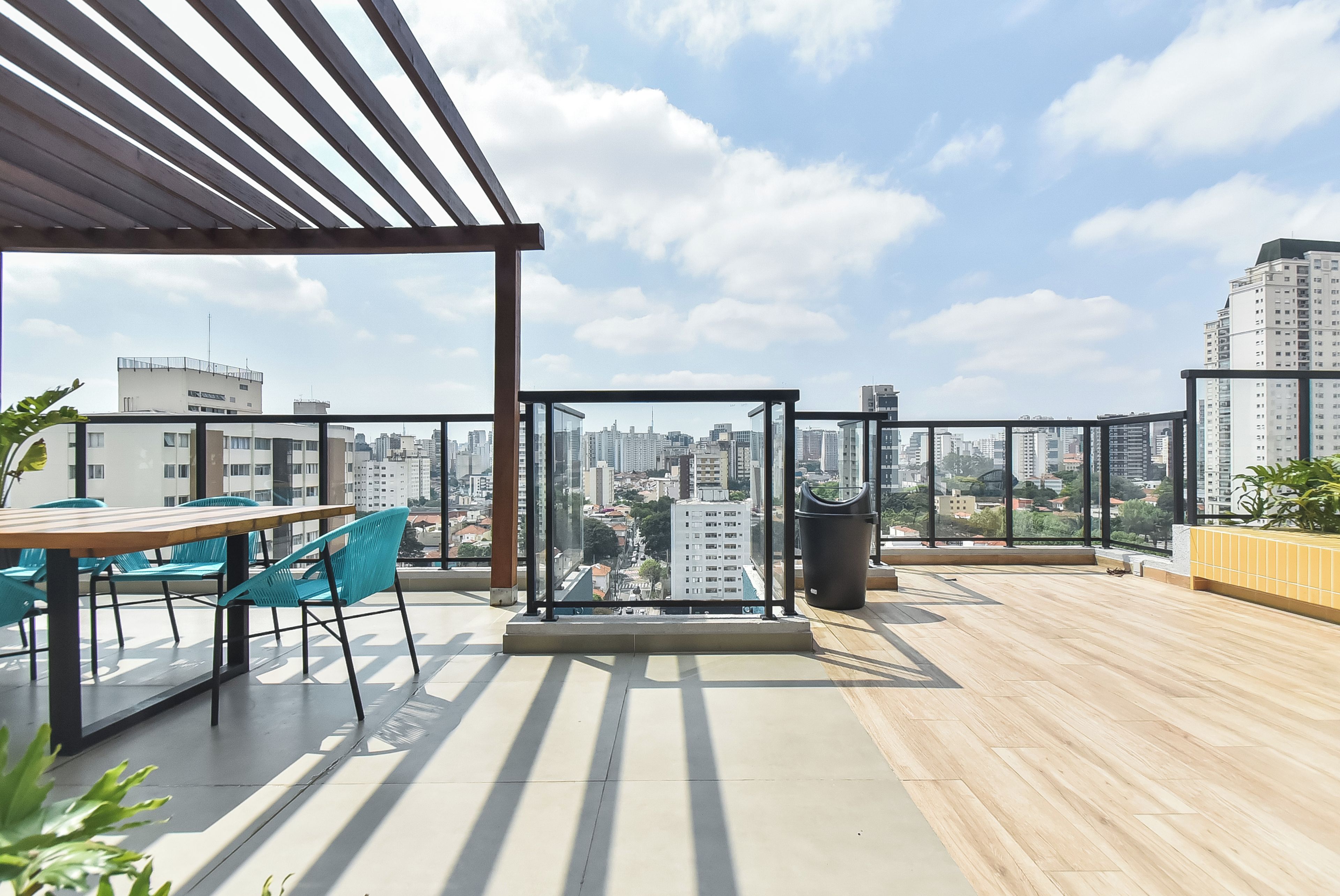 An outdoor terrace with a wooden floor, a table with chairs, a glass partition, and a view of a cityscape.