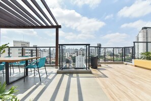 An outdoor terrace with a wooden floor, a table with chairs, a glass partition, and a view of a cityscape. - StayHome Vila Clementino Nurban - Pool  (São Paulo)