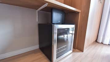 A compact refrigerator with a glass door, placed under a wooden shelf in a room with light-colored walls and flooring.