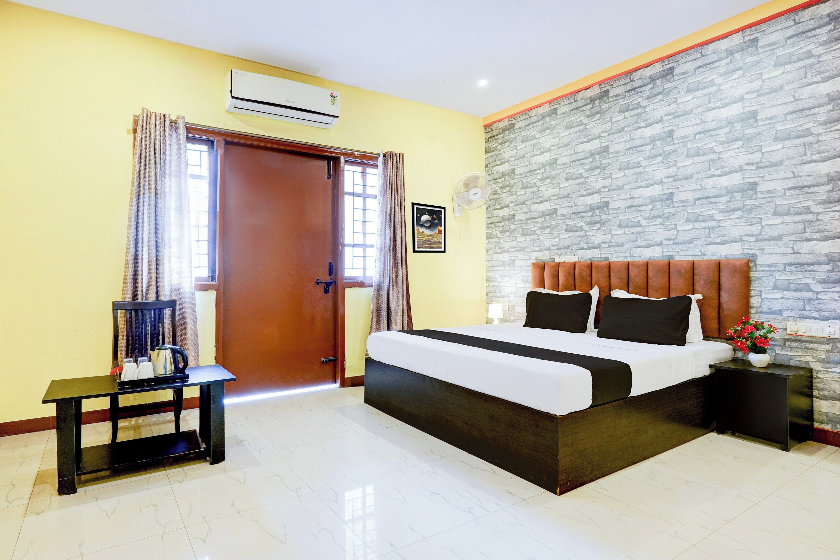 A hotel room with a bed, a nightstand, a table with a kettle, a wall-mounted air conditioner, and a stone-textured accent wall.
