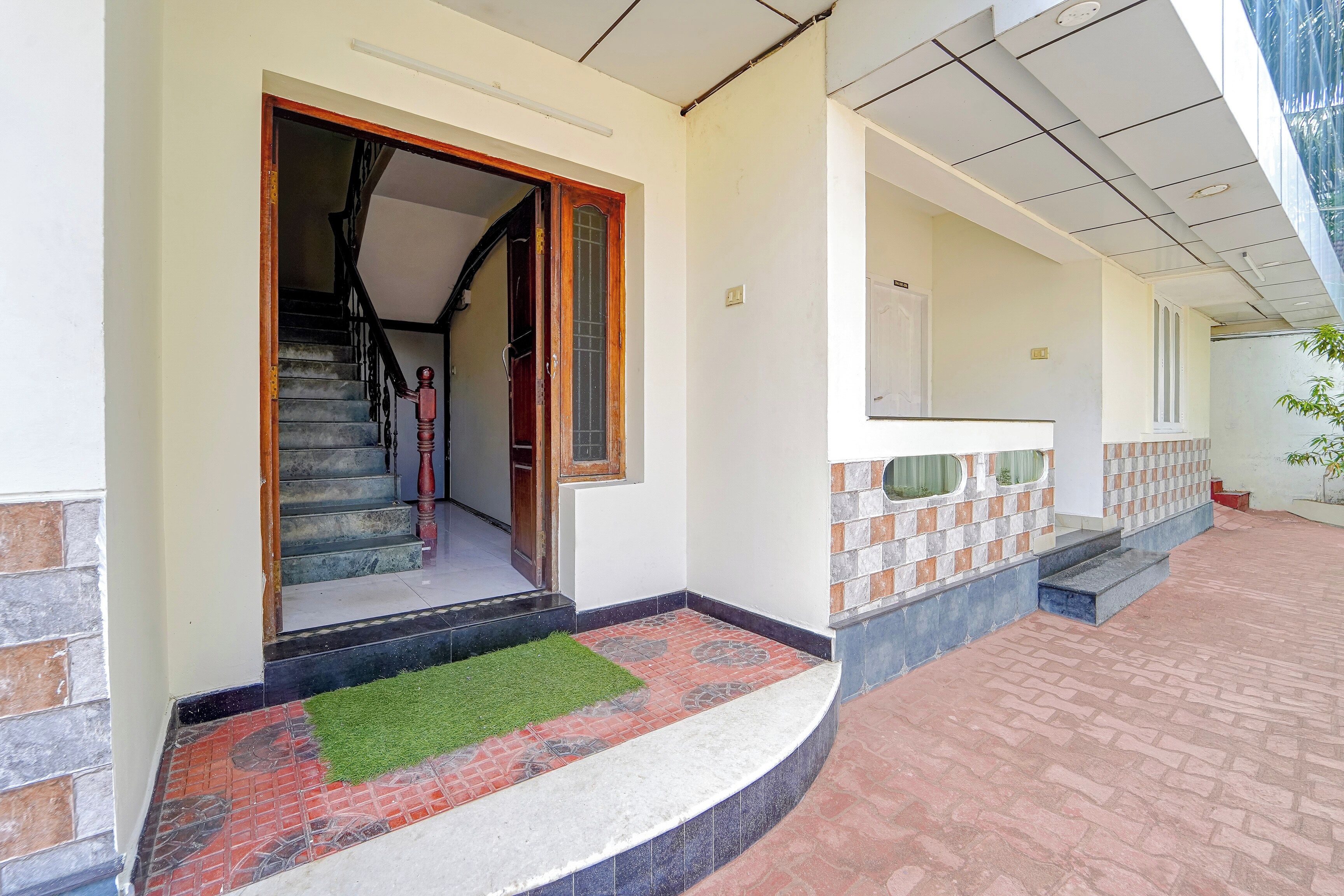A house entrance with a staircase leading to the interior, a small front yard with a green mat, and a tiled exterior wall.