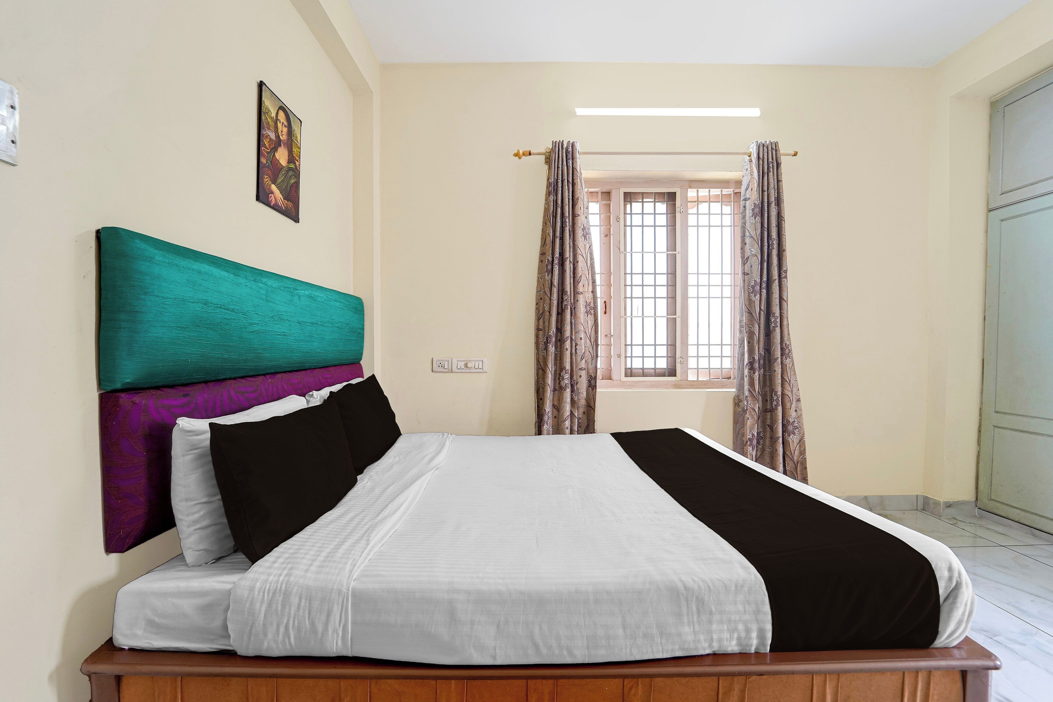 Un dormitorio con cama, cabecera, ventana con cortinas y una lámpara fijada a la pared.
