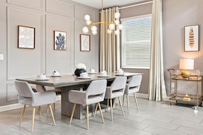 A dining area with a table set for six, six grey upholstered chairs with wooden legs, a chandelier with multiple light bulbs, and framed artwork on the walls. - 5-bedroom House in Championsgate With Private Pool (Kissimmee)