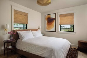 A bedroom with a large bed, two windows with blinds, a bedside table with a lamp, and a painting on the wall. - Stardust Ranch | The Ziggy House (Fayetteville)
