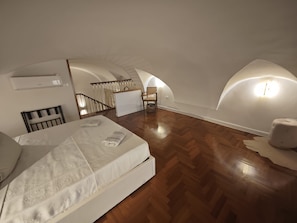 Ein Schlafzimmer mit Holzboden, einem Bett mit weißer Matratze, einem Sessel und einer Treppe, die zu einer oberen Ebene führt.