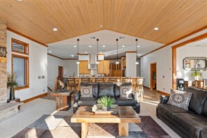 A spacious living area with a wooden ceiling, a central seating arrangement, and a kitchen in the background. - Stillwater Ranch - Mountain Ranch w/ River, Pond, Sauna, Stables! (Coalville)