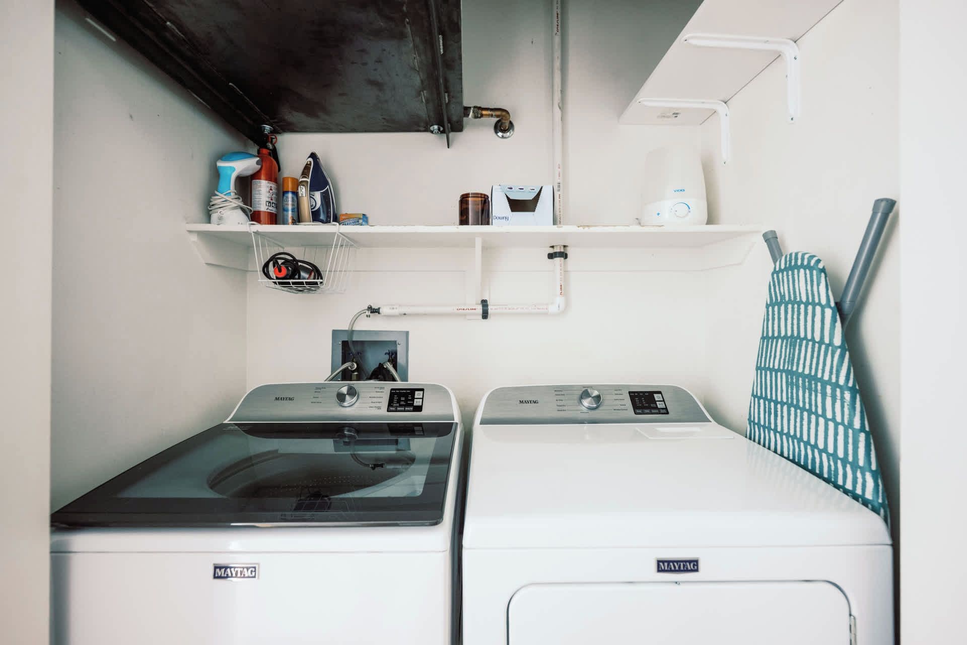 A laundry area with two Maytag washing machines, a shelf with various items, and a towel rack.