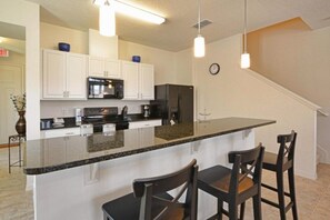 A modern kitchen with white cabinets, a black refrigerator, a microwave, and a granite countertop. There are two bar stools and a clock on the wall. - Disney 4BR Home w/ Pool & Lake View (Kissimmee)