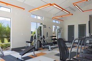 Academia com equipamentos de ginástica, grandes janelas e luminárias com iluminação laranja.