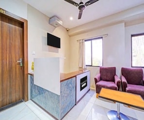 A hotel room with a reception desk, a ceiling fan, a TV mounted above the desk, two purple armchairs, a small table, and a window with curtains. - Deluxe Room Hotel Expression (Indore)