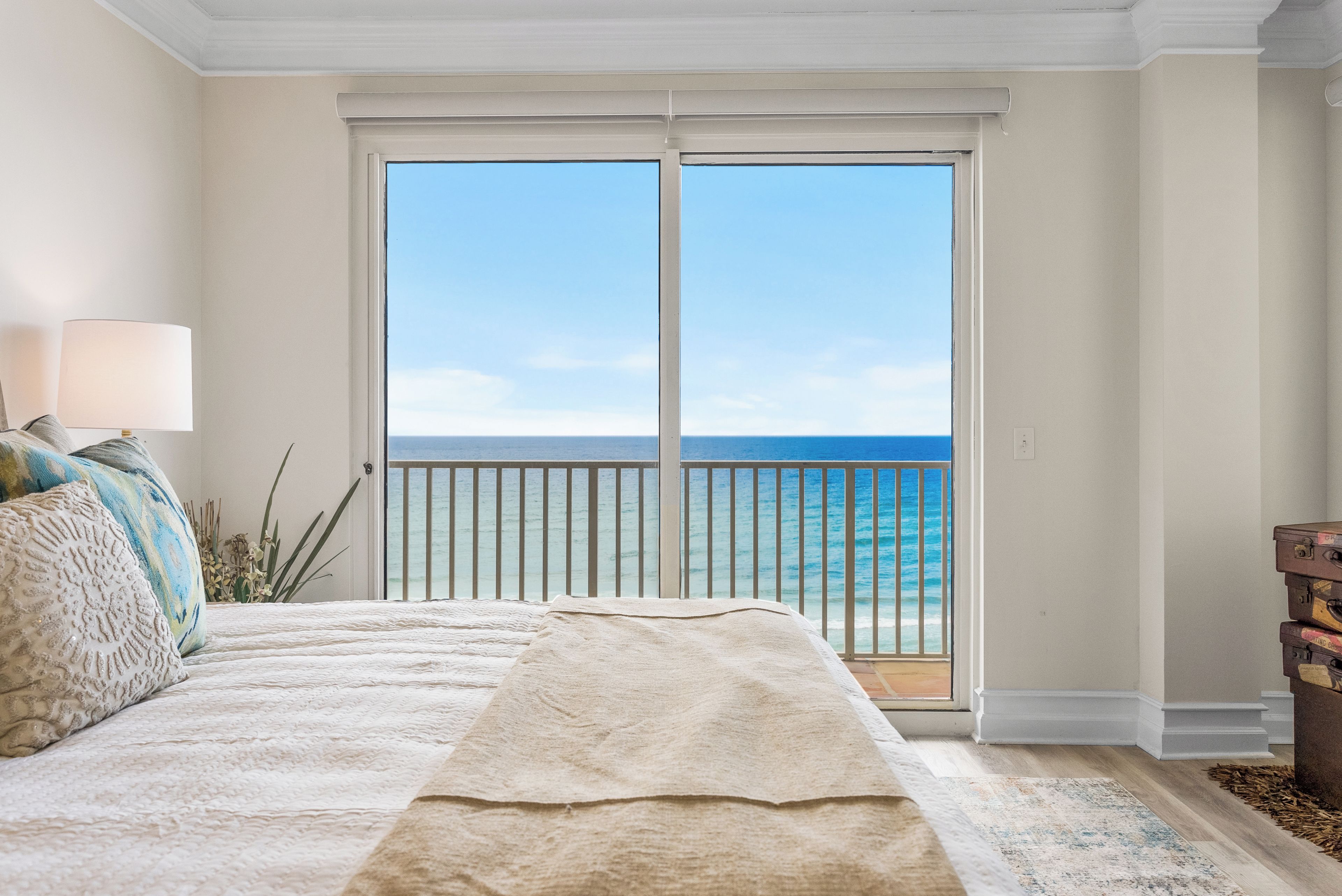 A bedroom with a large bed, a nightstand, a lamp, and a view of the ocean through a sliding door.