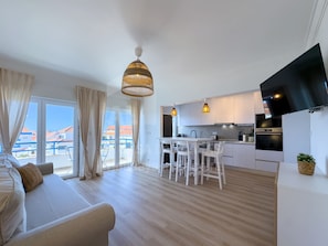 A modern kitchen with white cabinets, a dining area with a white table and chairs, and a large window with curtains. - Barbosa's House by ACasaDasCasas (Ericeira)