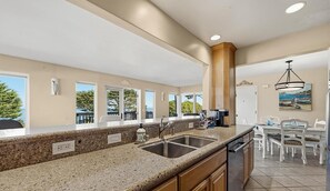 A kitchen with granite countertops, a double sink, and a dining area with white chairs and a table. - Anchors Away - Pacific Grove Condo - Your Oceanfront Beach Pad (Pacific Grove)