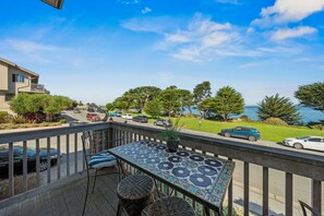 Een balkon met een tafel en stoelen, uitkijkend op een straat met geparkeerde auto's en een uitzicht op de zee.