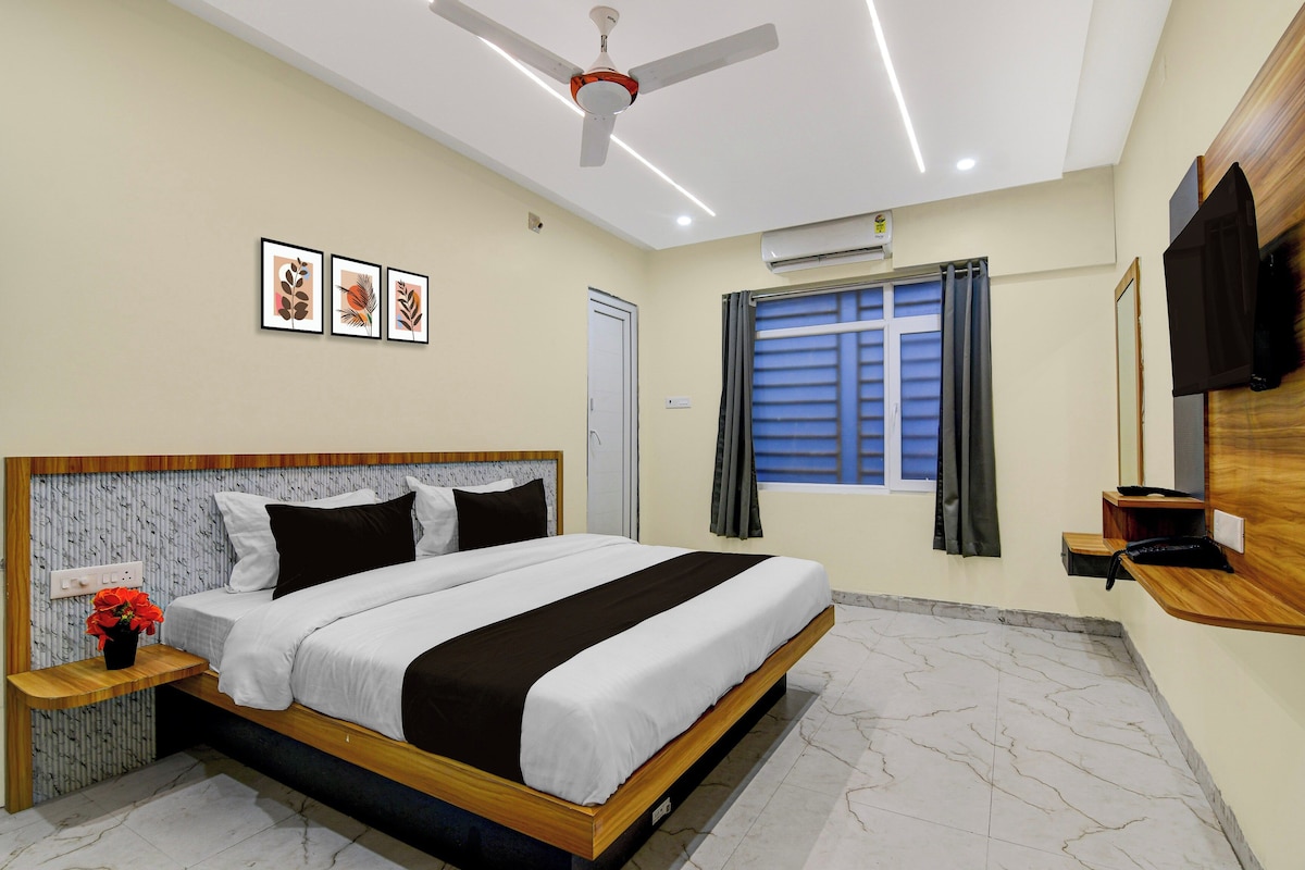 A hotel room with a bed, a ceiling fan, framed artwork, an air conditioning unit, a window with curtains, and a wooden nightstand with a vase of flowers.