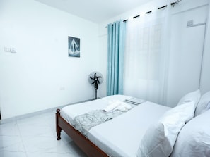 Ein Schlafzimmer mit einem Bett, einem Standventilator, einem Fenster mit Vorhängen und einem an der Wand montierten Lichtschalter.