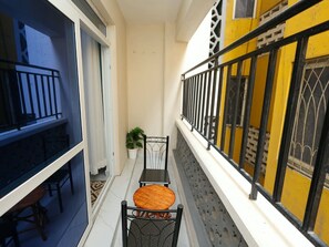 Ein schmaler Balkon mit Tisch und Stühlen, einem Blumentopf und einer gelben Wand mit schwarzem Geländer.