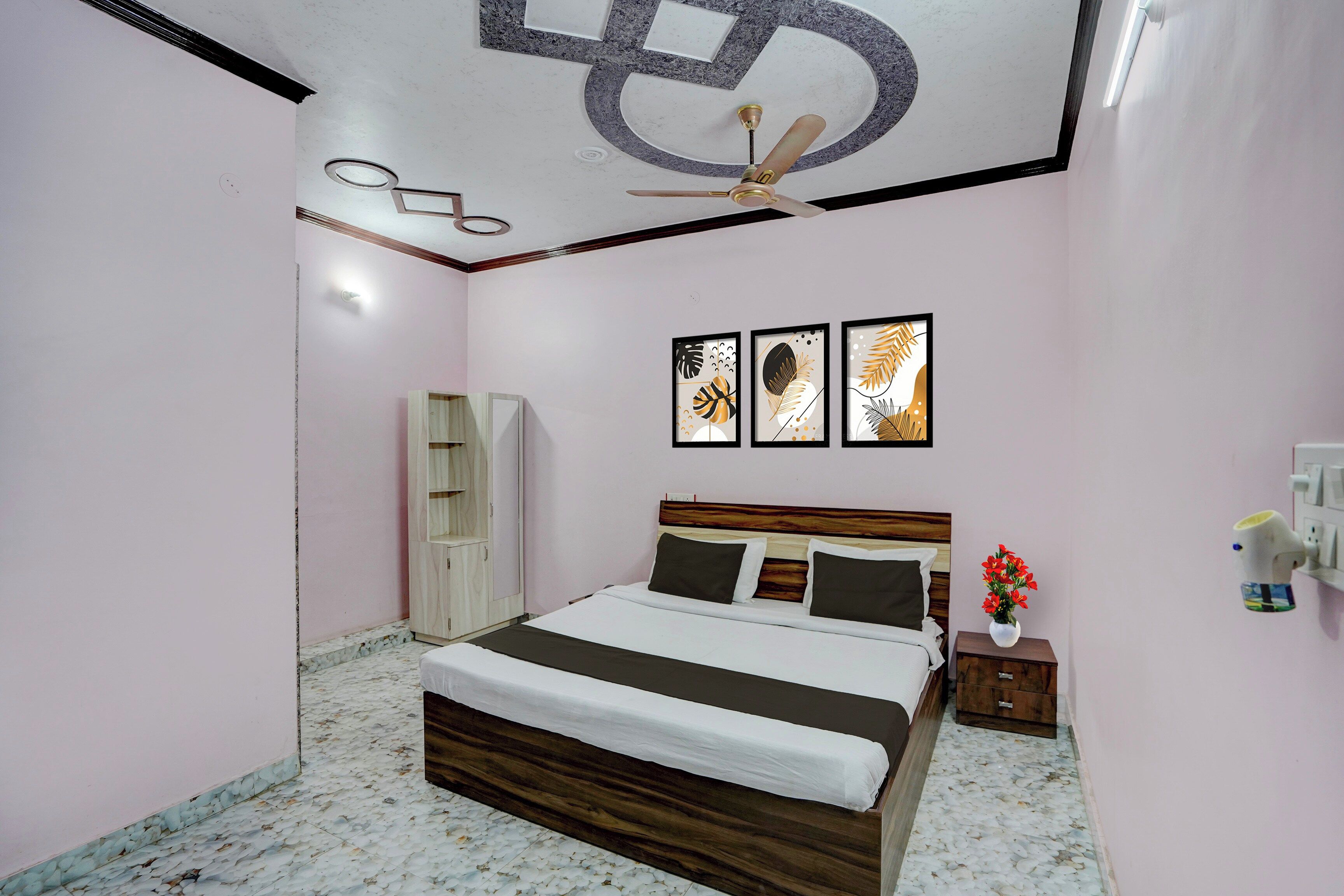 A bedroom with a bed, a ceiling fan, a nightstand, a vase with flowers, and framed artwork on the wall.