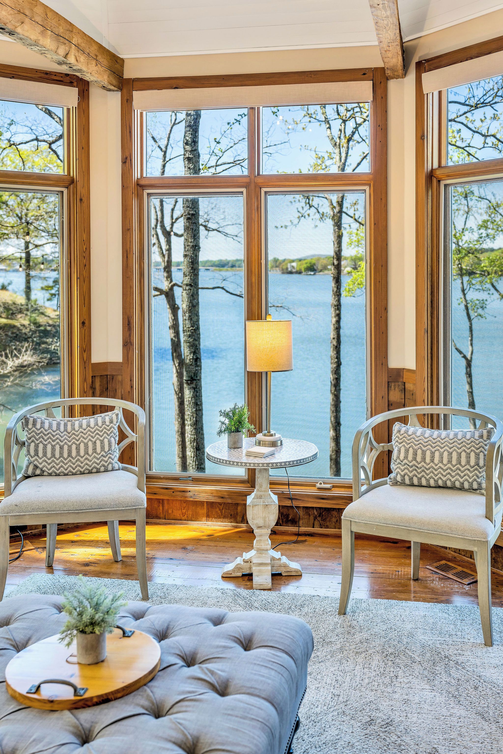 A room with a view of a lake, wooden beams, two upholstered chairs, a round table with a lamp, and a tufted ottoman.