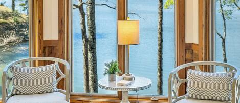 A room with a view of a lake, wooden beams, two upholstered chairs, a round table with a lamp, and a tufted ottoman.