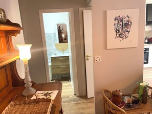 Ein Zimmer mit einem Holztisch, einer Lampe, einem Korbwannenstuhl, einer Tür zu einem weiteren Raum und einem an der Wand hängenden Kunstwerk.
