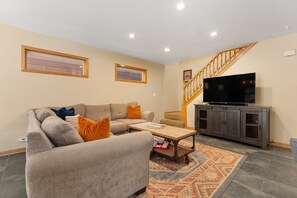 A living room with a sofa, coffee table, TV, and a staircase. - Spacious 5BR Wildernest Home | Private Hot Tub & Views (Silverthorne)