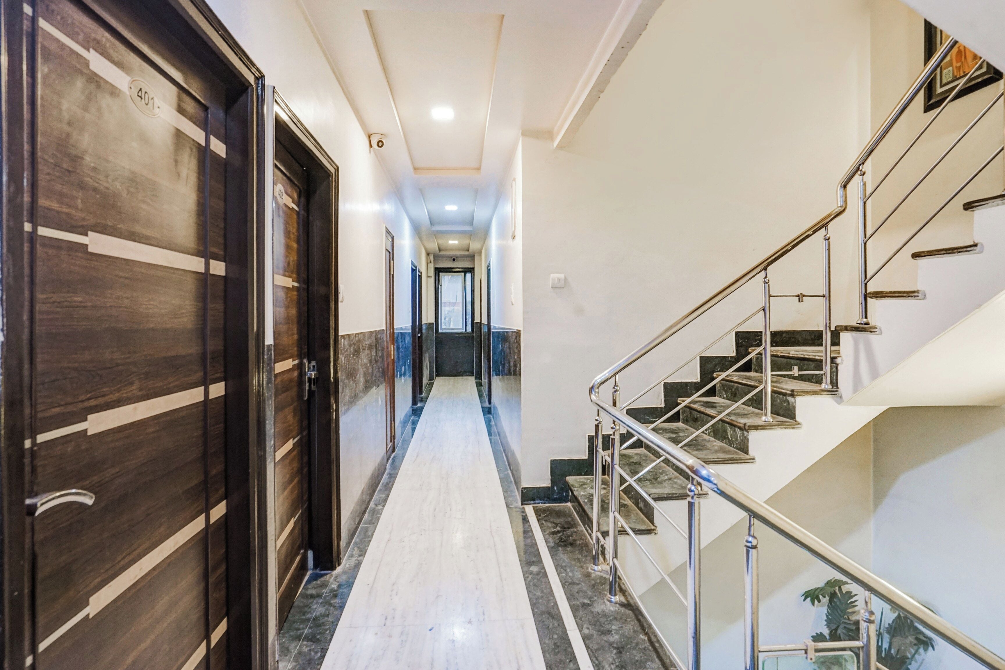 Un pasillo de hotel con piso de mármol, una escalera con barandillas de metal y puertas de madera con placa de número.