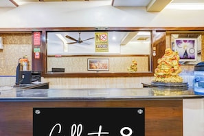 Una recepción de hotel con un cartel de circuito cerrado de televisión, un letrero de 'no se permite comer fuera', un ventilador de techo y una estatua.