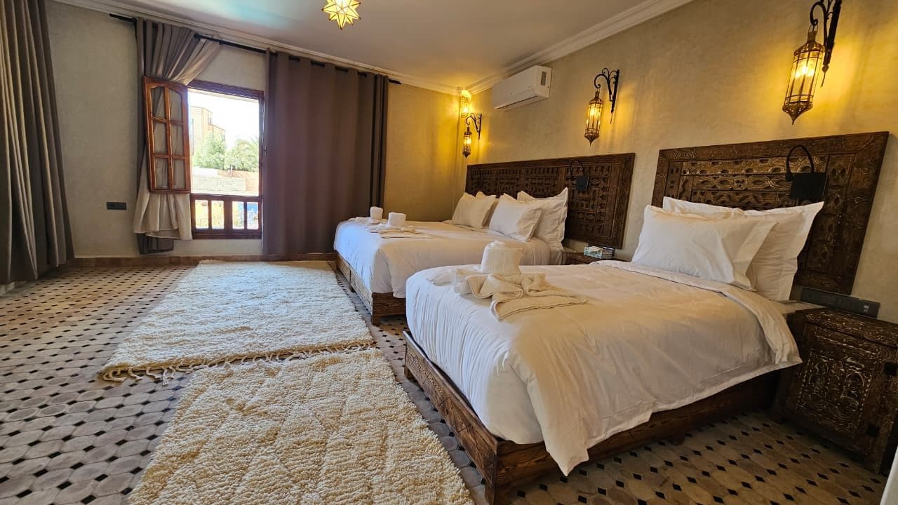 Une chambre d’hôtel avec deux lits, une grande fenêtre avec des rideaux, un sol à motifs et des lampes fixées au mur.