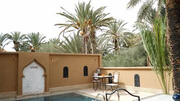 Zona junto a la piscina con una mesa y sillas, rodeada de palmeras y una pared color arena con una puerta.