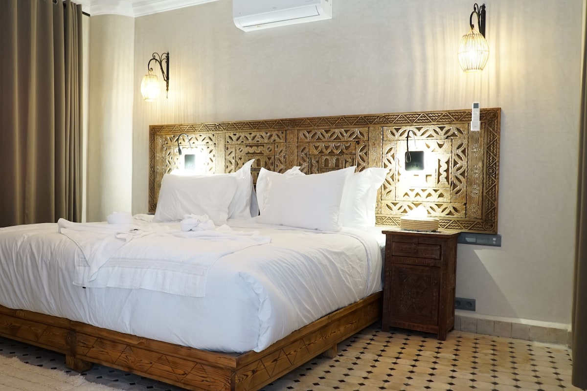 Habitación de hotel con cama de madera, ropa de cama blanca, cabecero estampado y dos lámparas fijadas a la pared.