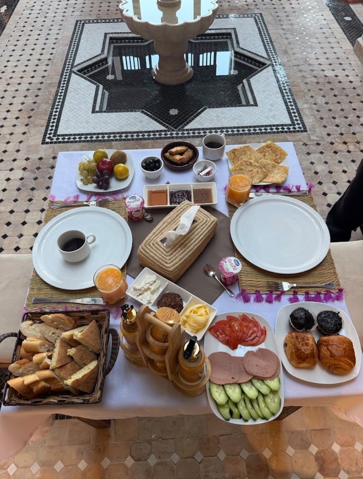 A table set with a variety of foods and drinks, including bread, fruits, and condiments, on a tiled floor with a decorative pattern.