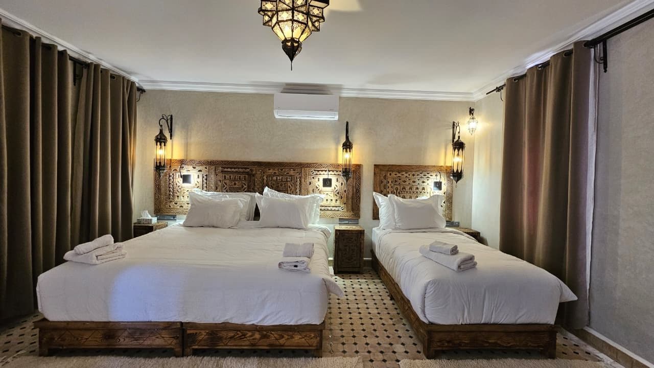 A hotel room with two beds, a patterned headboard, wall-mounted lamps, and a ceiling light fixture.