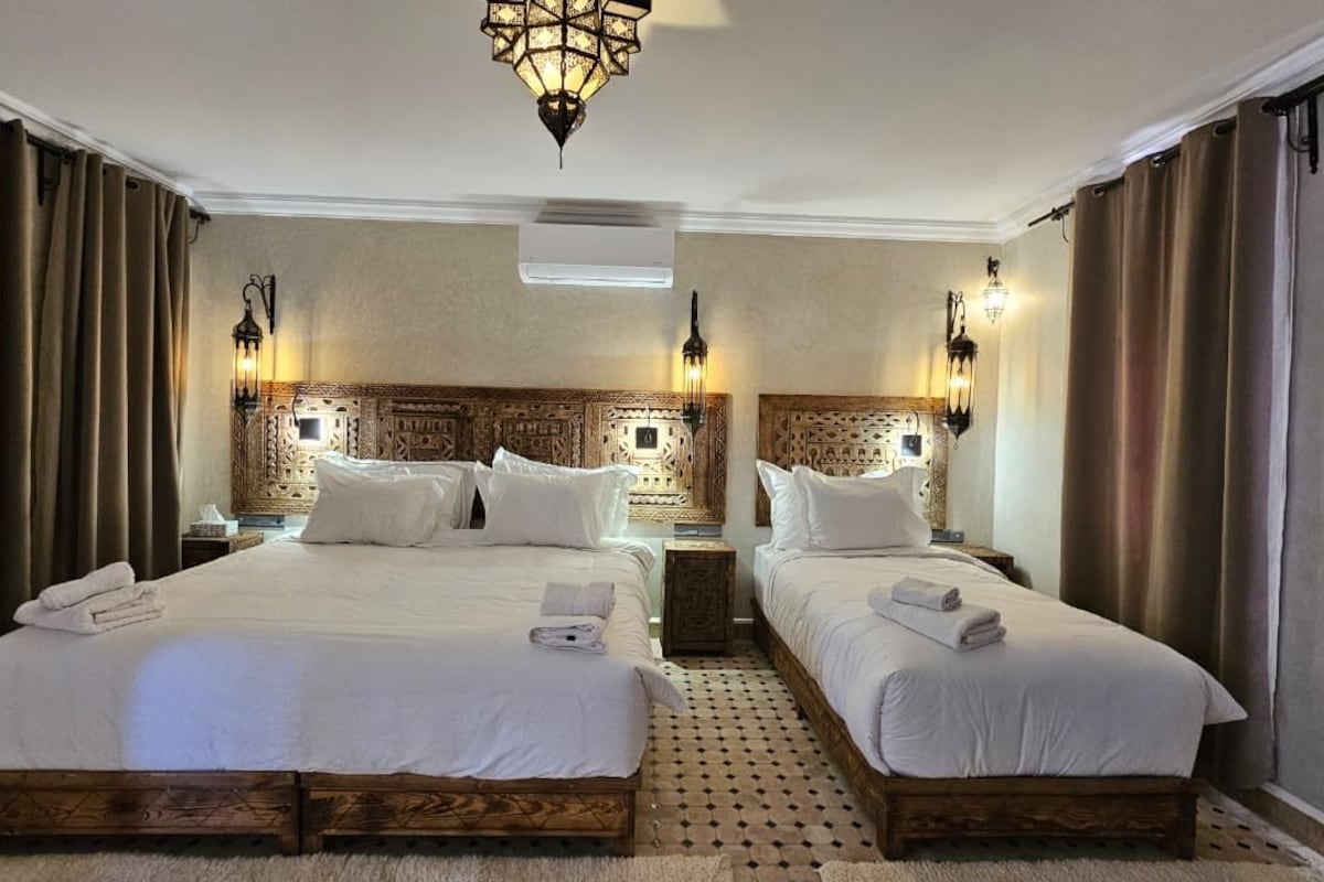 A hotel room with two beds, a patterned headboard, wall-mounted lamps, and a ceiling light fixture.