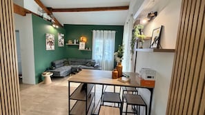 A modern living space with a green accent wall, a grey sectional sofa, a wooden dining table with four chairs, and a kitchen area with appliances and storage. - New Listing (Nîmes)