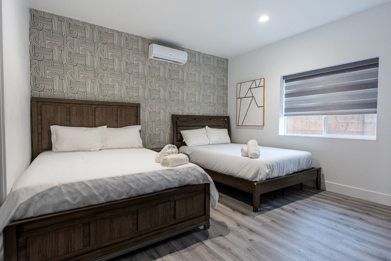 Habitación de hotel con dos camas, papel tapiz estampado, una ventana con persianas y un aire acondicionado de pared.