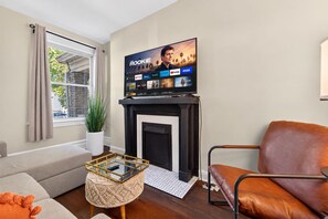 Ein Wohnzimmer mit Kamin, Fernseher der einen Film zeigt, einer Couch, einem Couchtisch und einer Zimmerpflanze.