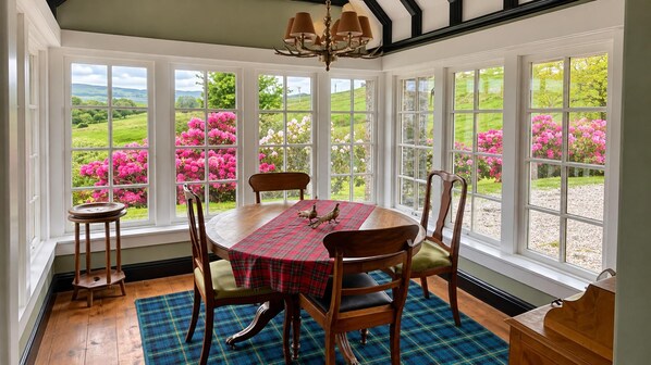 A dining area with a table set for four, surrounded by chairs, with a view of a garden through large windows.