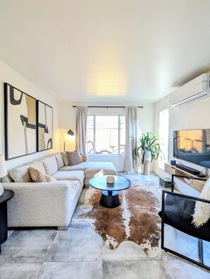 A modern living room with a sectional sofa, a round coffee table, a television, and a large window with curtains. - Downtown Kelowna Carriage House | Private Hot Tub, Firepit & Fenced Yard (Kelowna)