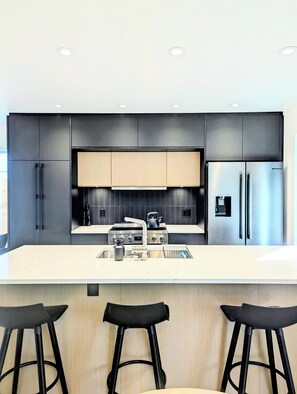 A modern kitchen with a central island, stainless steel appliances, and black bar stools. - Downtown Kelowna Carriage House | Private Hot Tub, Firepit & Fenced Yard (Kelowna)