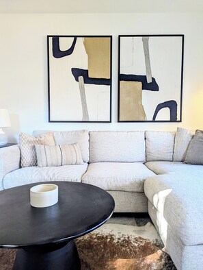 A living room with a sectional sofa, a round coffee table, and two abstract paintings on the wall. - Downtown Kelowna Carriage House | Private Hot Tub, Firepit & Fenced Yard (Kelowna)