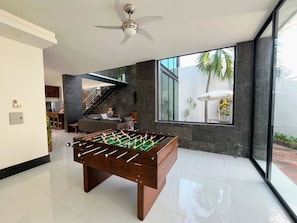 A modern living room with a foosball table, a ceiling fan, and a large window offering a view of the outdoors. - 4-king size beds bedroom- house in peaceful Sayulita walk to the beach (Sayulita)