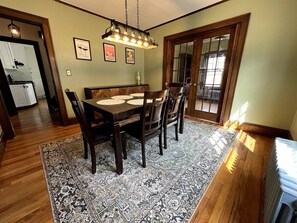 Uma sala de jantar com mesa e cadeiras de madeira, tapete estampado e luminária pendente.