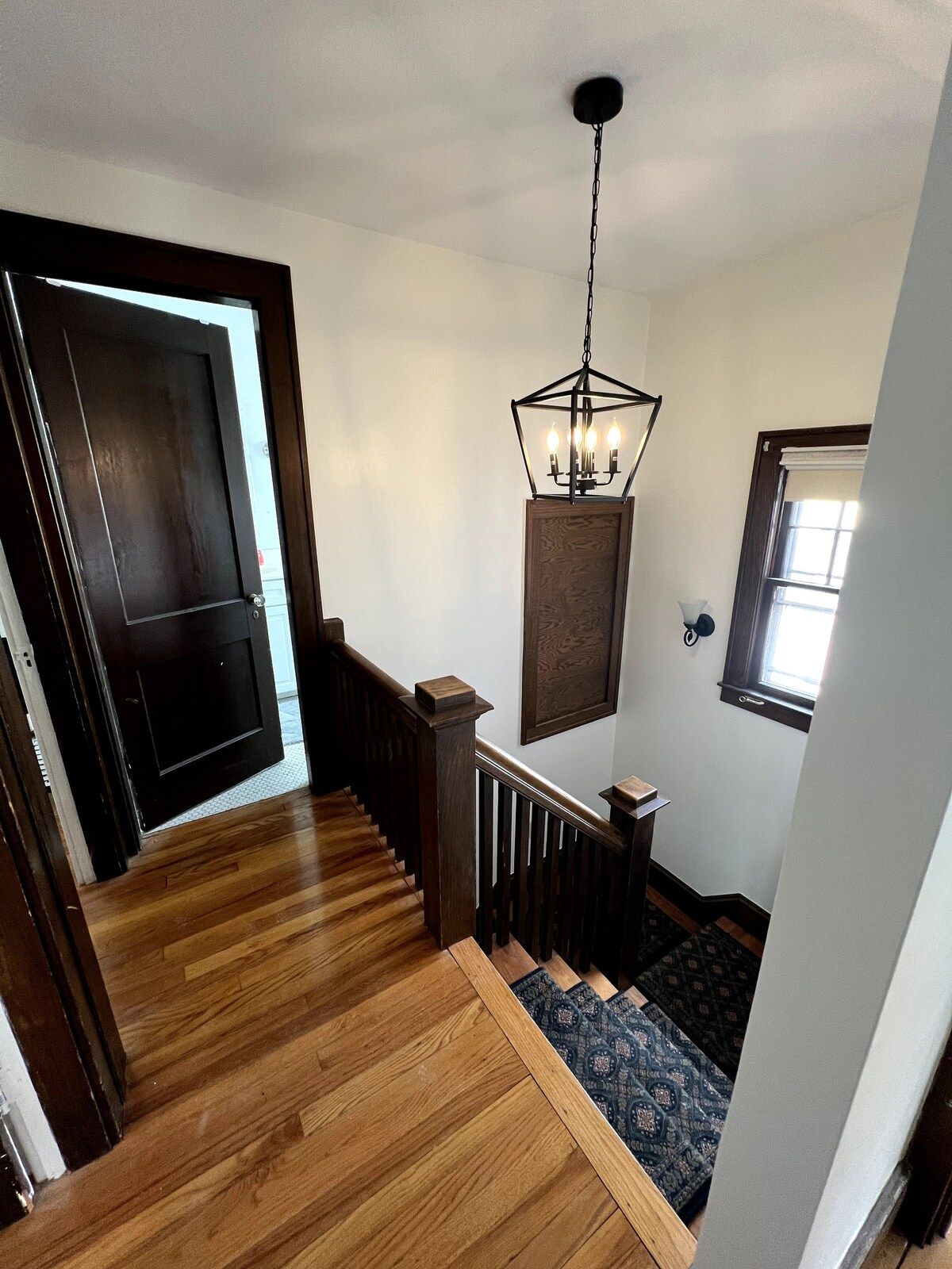 A wooden staircase with a dark railing, a hanging light fixture, and a window with a white blind.