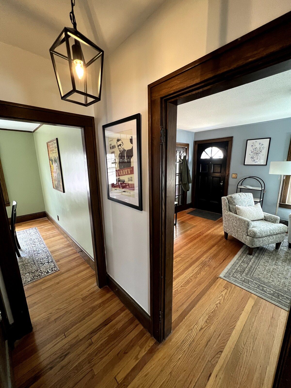A hallway with wooden flooring, a hanging light fixture, and a door leading to a room with a patterned rug and a chair.