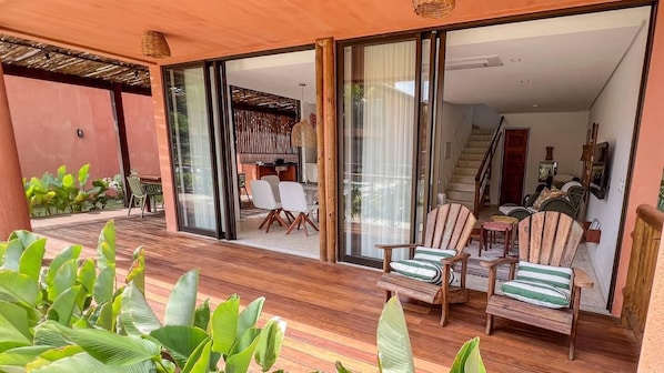 Un espacio exterior cubierto con una terraza de madera, asientos y puertas correderas de vidrio que dan a un ambiente interior con una escalera y un comedor.