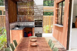 An outdoor kitchen with a grill, a dining table with chairs, and a view of the surrounding garden.