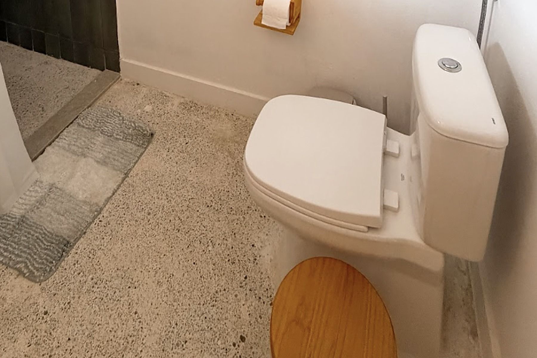 Un inodoro con tanque y asiento blancos, un cuenco de madera y un piso beige moteado.