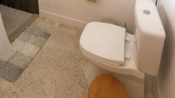 Un inodoro con tanque y asiento blancos, un cuenco de madera y un piso beige moteado.