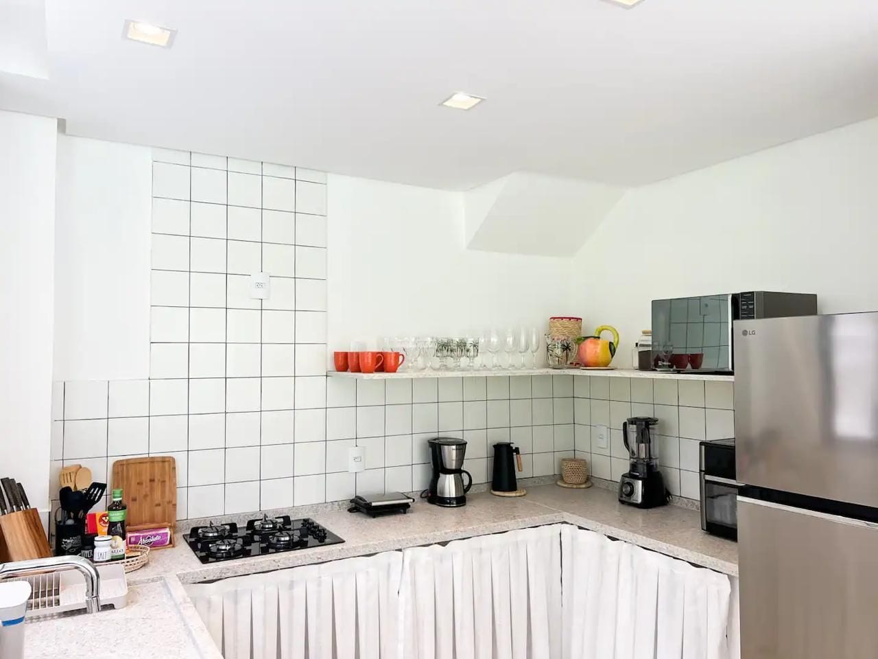 Una cocina moderna con paredes de azulejos blancos, un refrigerador de acero inoxidable y varios electrodomésticos como una cafetera y una licuadora.